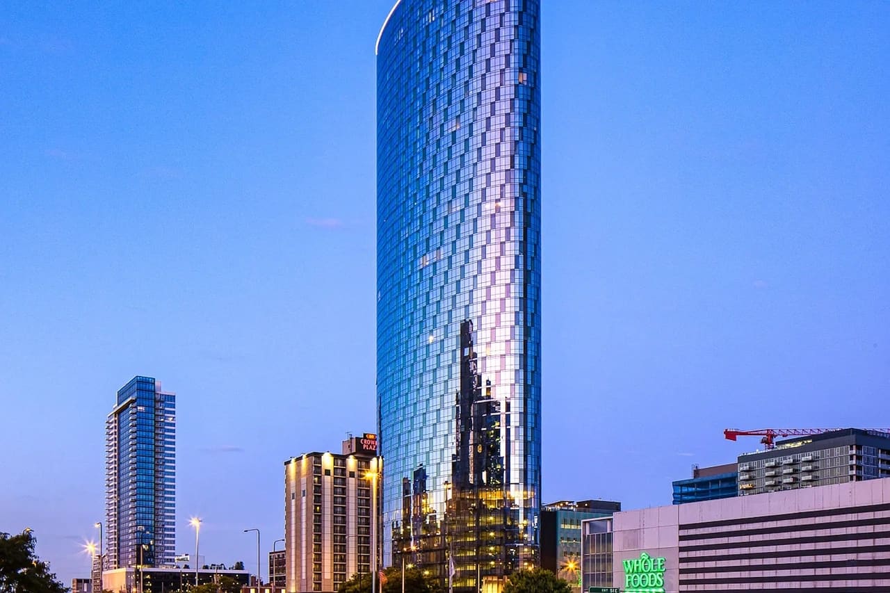 Tall cylindrical glass tower with blue and purple facade at dusk, surrounded by other downtown buildings and city lights.
