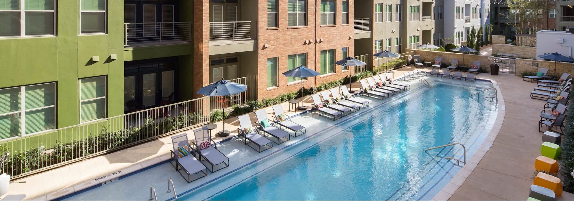 Curved outdoor swimming pool with lounge chairs, umbrellas, and multi-story brick and green apartment buildings.