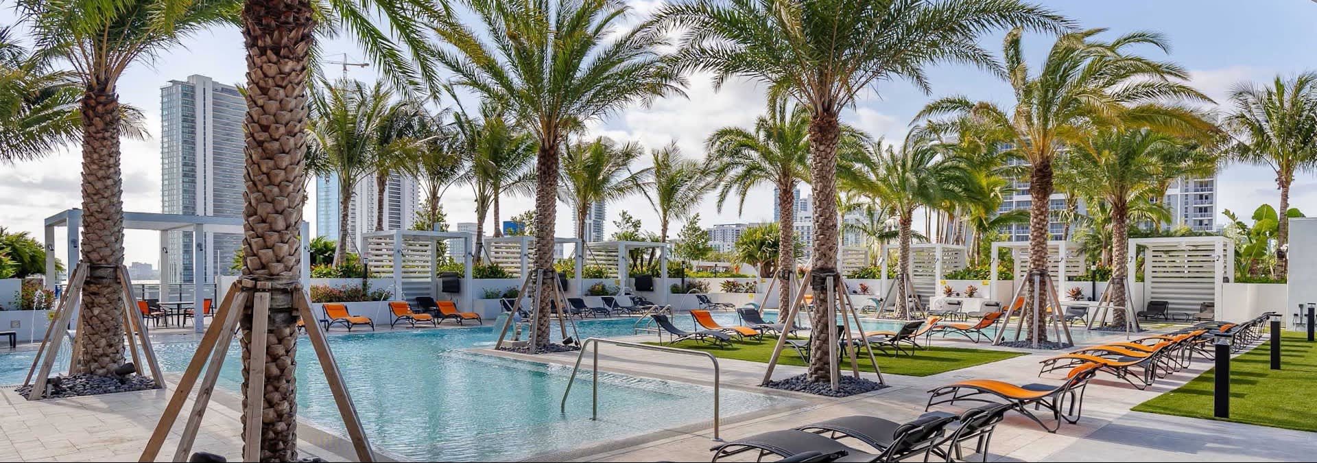 Resort pool with turquoise water, palm trees, lounge chairs, and Miami skyline in background.