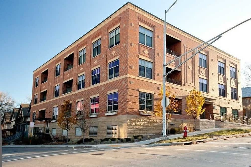 Four-story brick apartment building with large windows, balconies, and stone foundation on a tree-lined street.