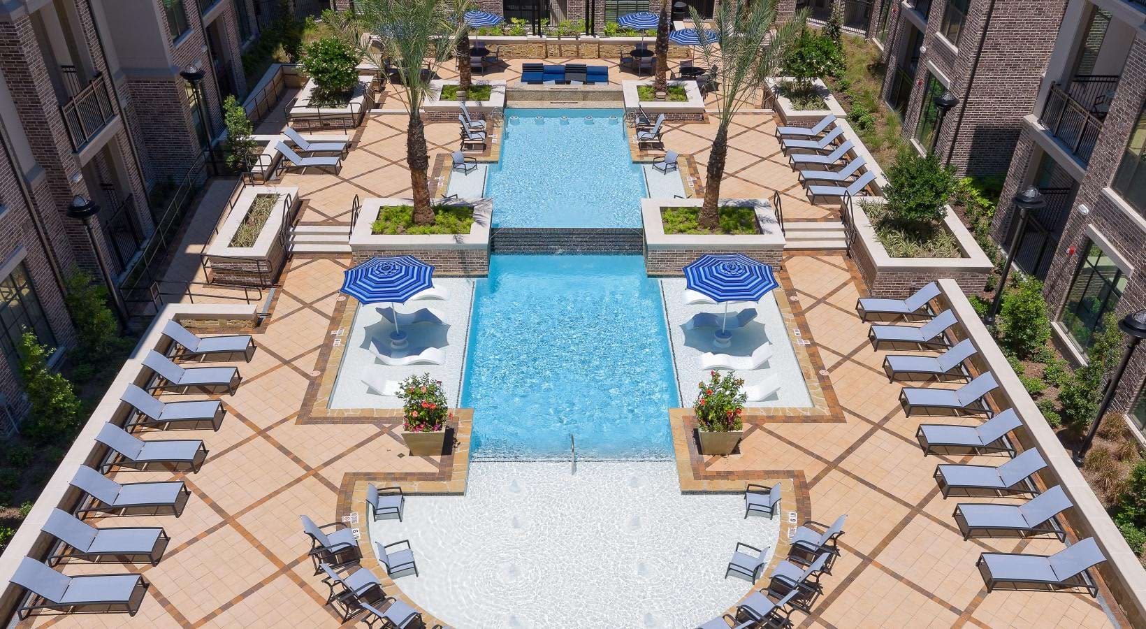 Aerial view of resort-style pool courtyard with blue umbrellas, lounge chairs, and tiered pools flanked by brick apartment buildings.