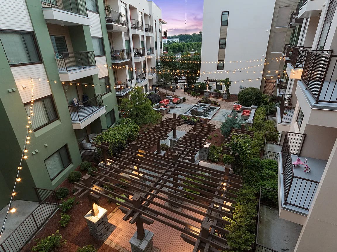 Elevated view of apartment courtyard with dark wooden pergolas, string lights, red seating, landscaping, and surrounding multi-story residential buildings at dusk.