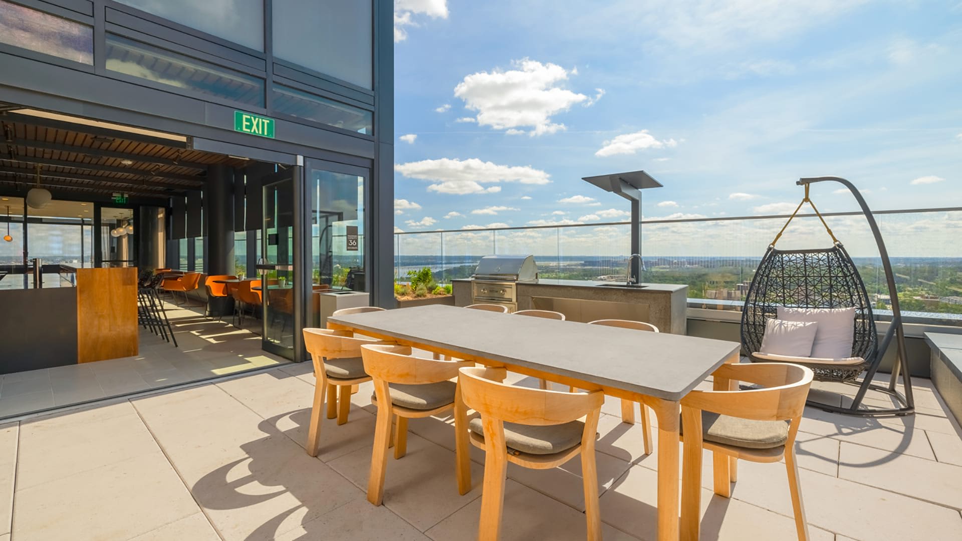 Rooftop terrace with wooden dining table, chairs, hanging egg chair, built-in grill, and panoramic city skyline views under blue sky.