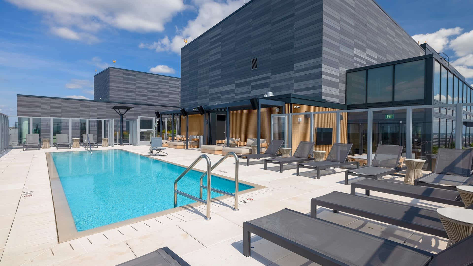 Outdoor pool with metal handrails surrounded by gray lounge chairs, white concrete deck, and multi-story gray residential buildings under blue sky.