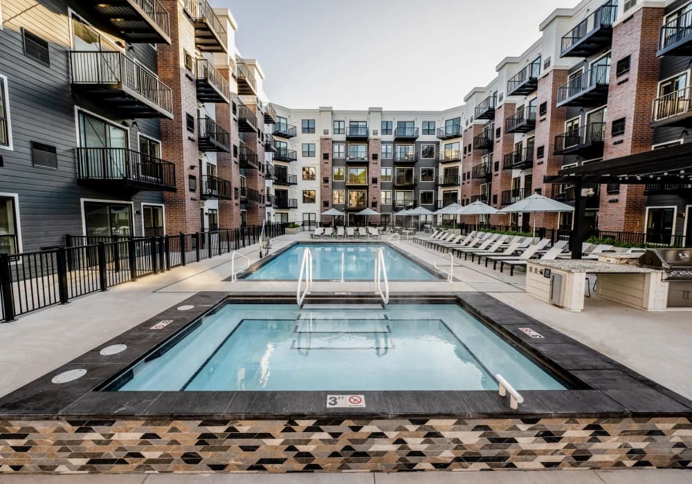 Two rectangular pools with white railings in a courtyard surrounded by multi-story brick apartment buildings and white umbrellas.