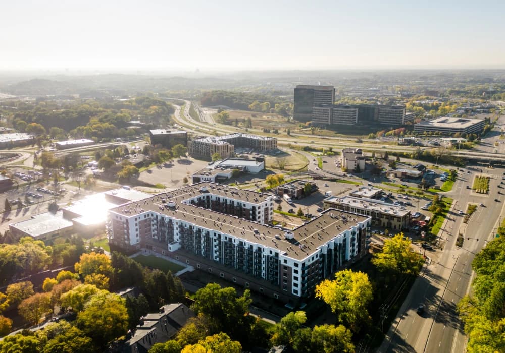Aerial view of a multi-story residential building with brick and blue facade, surrounded by green trees, parking areas, and a highway in the distance.