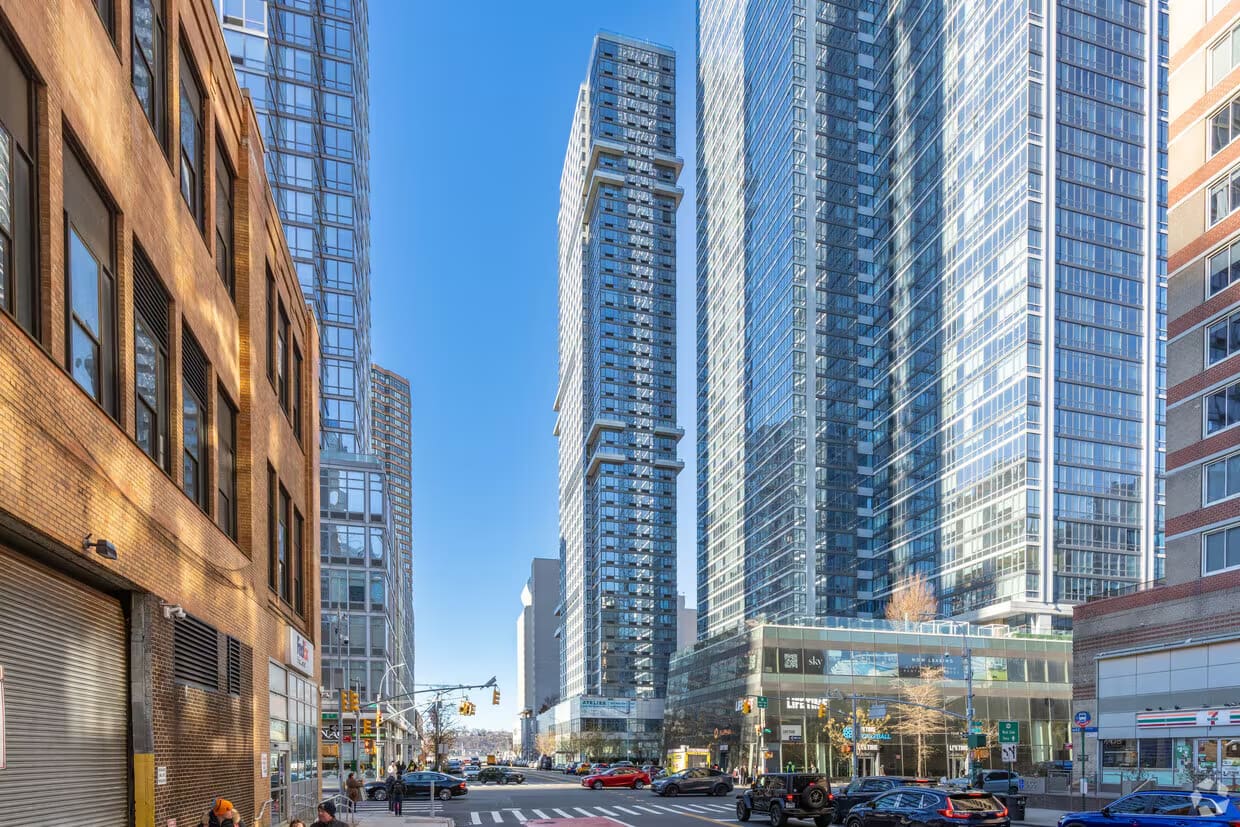 Urban street view of tall residential buildings, storefronts, and pedestrians under clear blue sky in New York City.