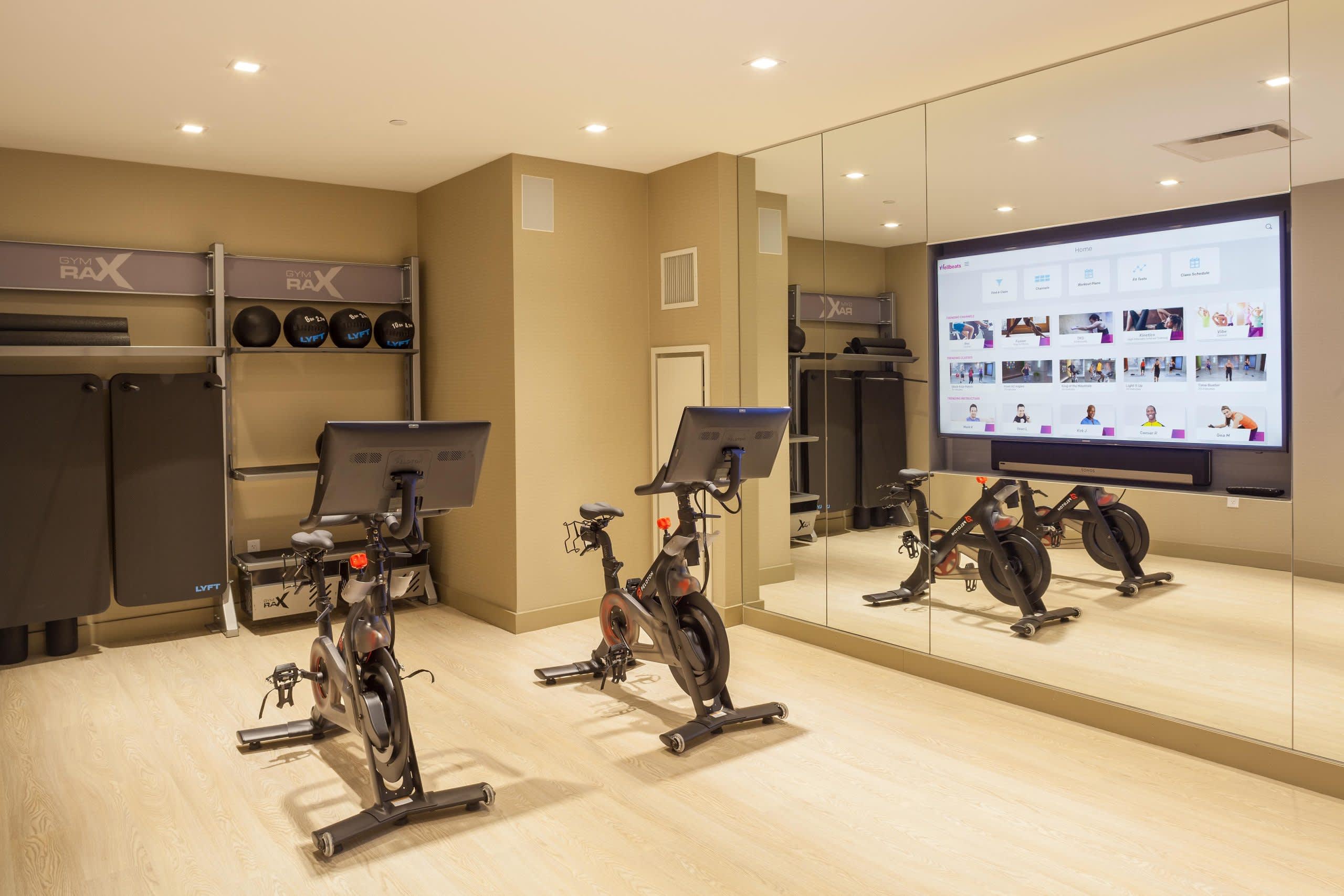 Modern fitness studio with three stationary bikes, mirrored walls, large digital screen, and equipment storage along beige walls.