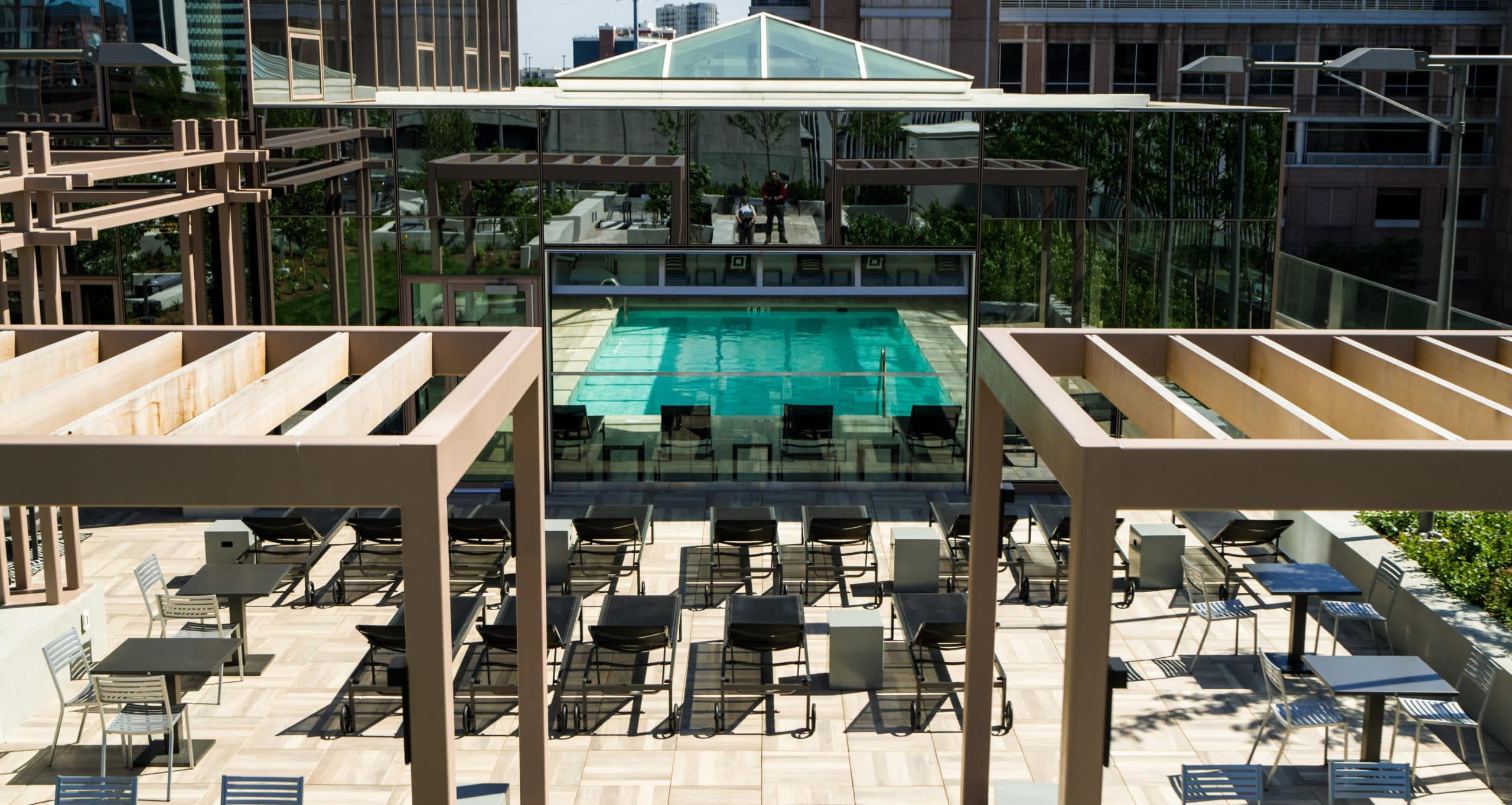 Rooftop terrace with wooden pergolas, lounge chairs, and view of indoor heated swimming pool with glass enclosure and downtown buildings beyond.