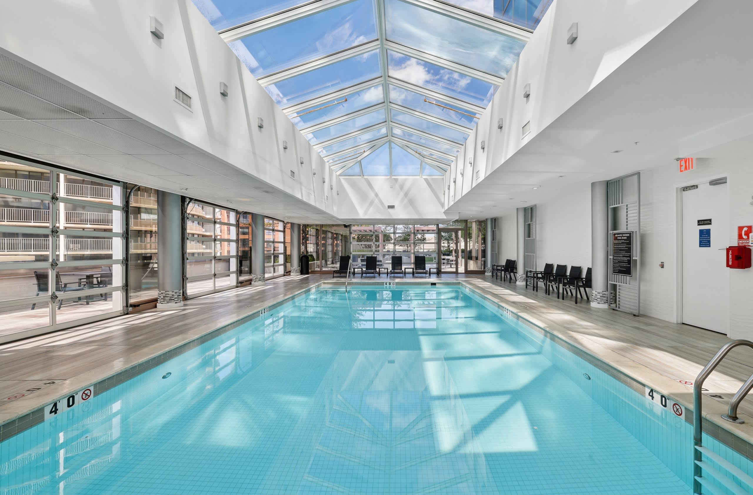 Indoor lap pool with clear blue water, white walls, skylights, and large windows showing urban views outside.