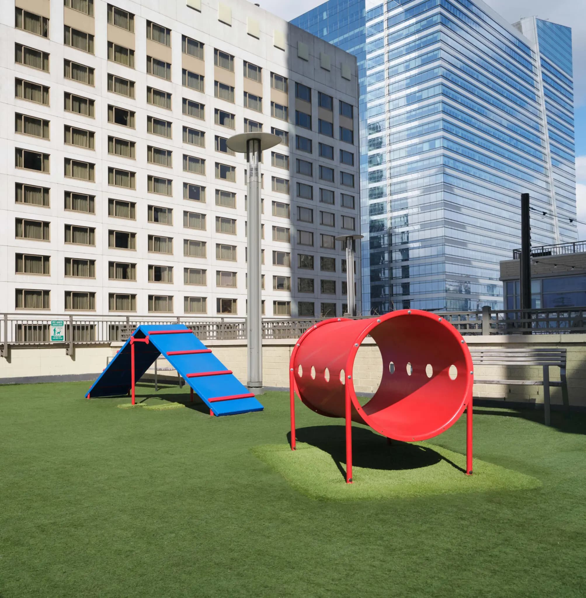 Rooftop play area with red tunnel climber and blue climbing structure on artificial turf, surrounded by residential and office buildings.