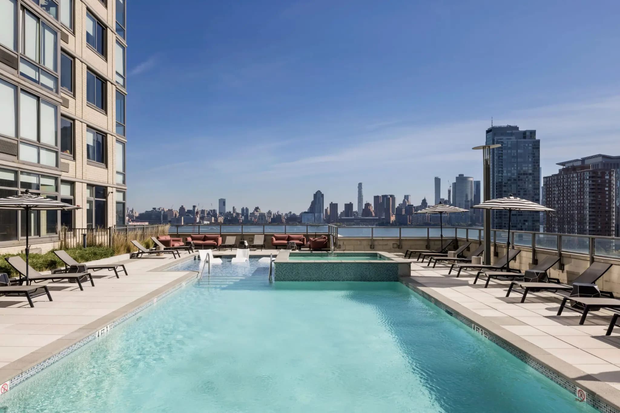 Rooftop pool with clear turquoise water, lounge chairs, umbrellas, and Manhattan skyline across the water.