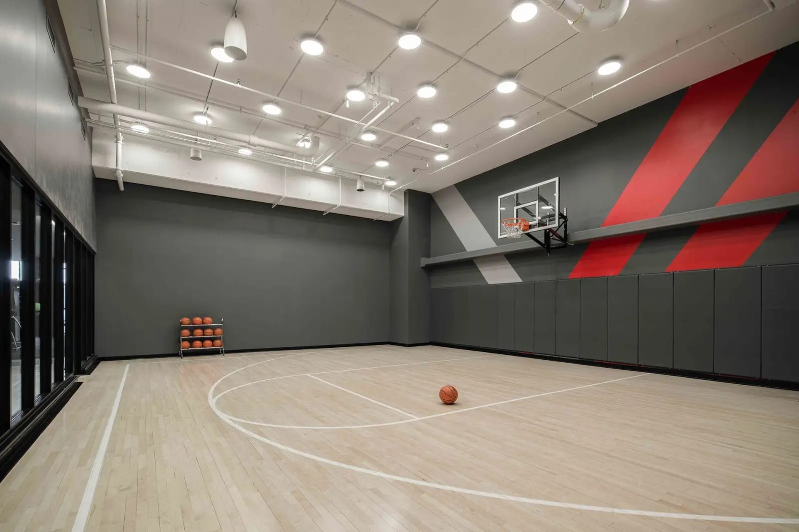 Indoor basketball court with wooden floor, white court markings, basketball hoop, red and gray geometric wall design, and basketball storage rack.