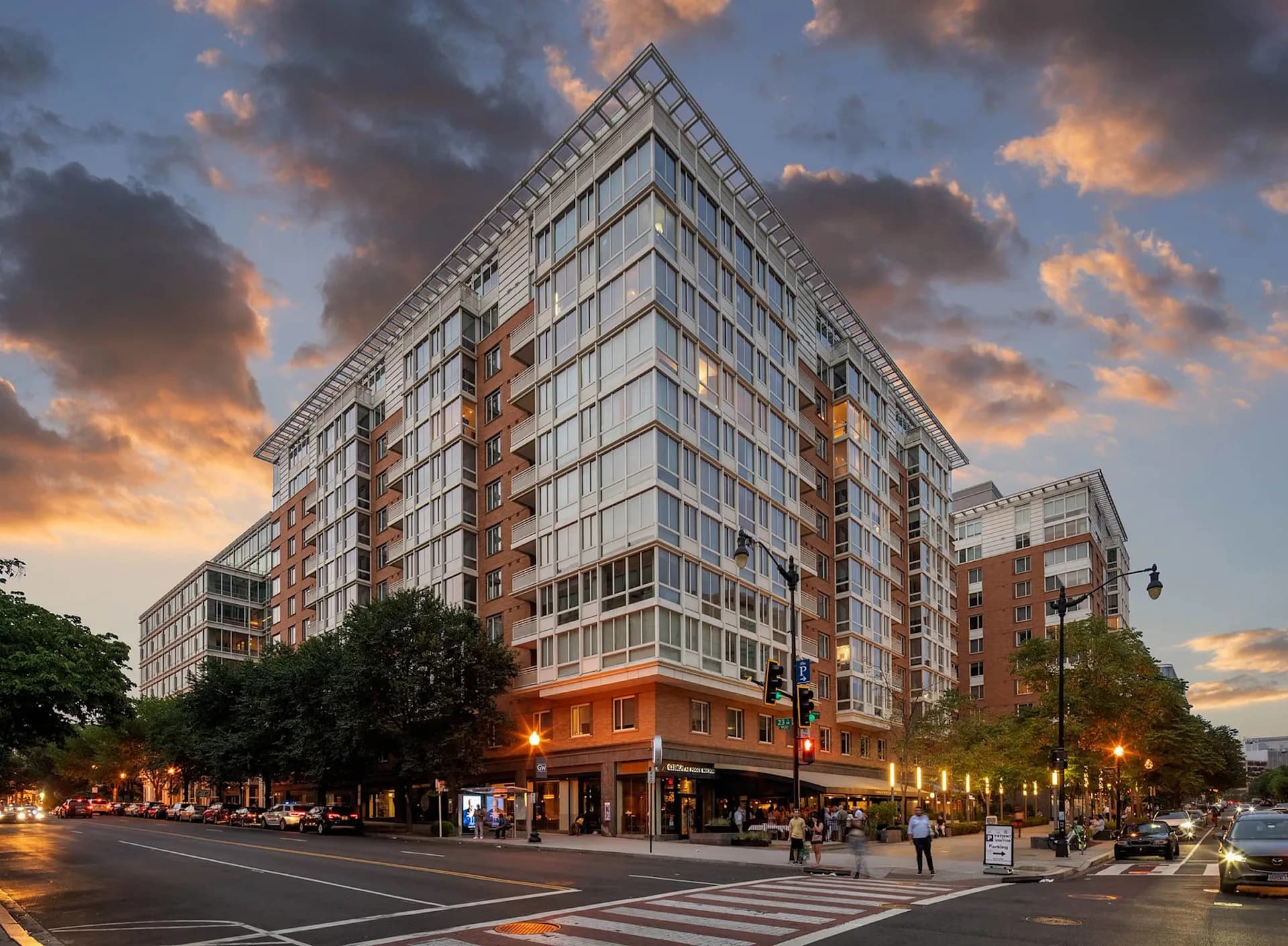 Corner view of mixed-use residential building with red brick base, upper floors of glass and metal, ground-level storefronts, and evening street lighting.