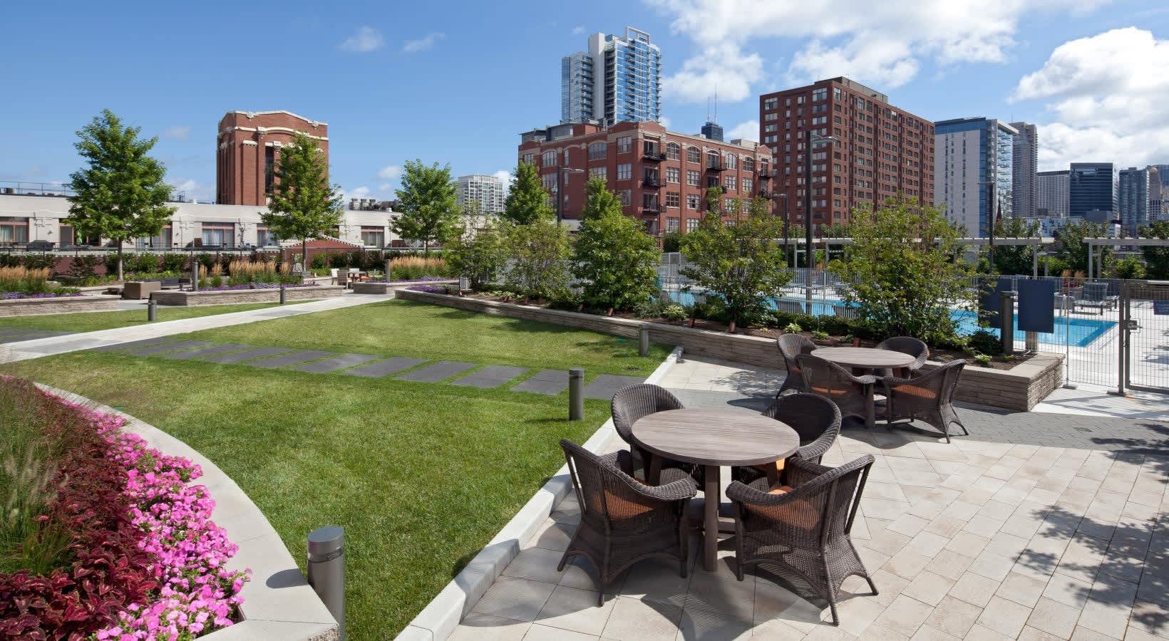 Rooftop terrace with round dining tables, wicker chairs, pink flowering shrubs, green lawn, and Chicago skyline backdrop.