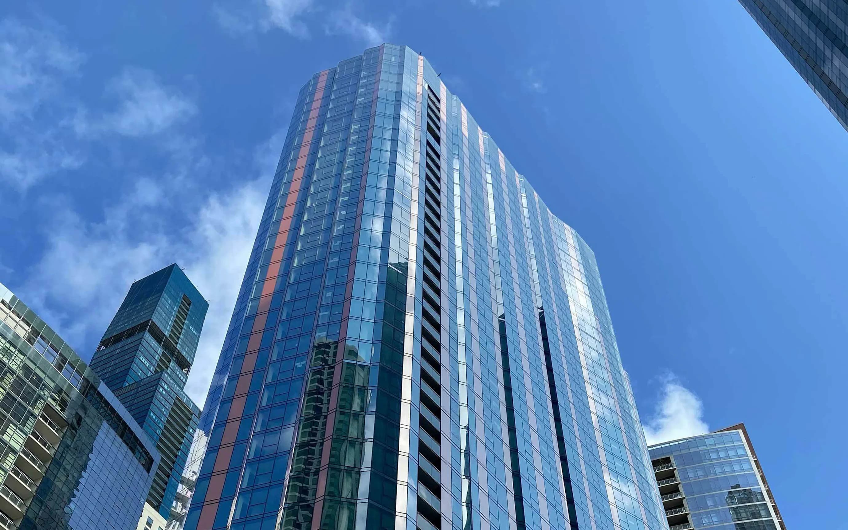 Tall residential tower with blue glass and metal facade, red accent stripe, surrounded by other downtown buildings under clear sky.