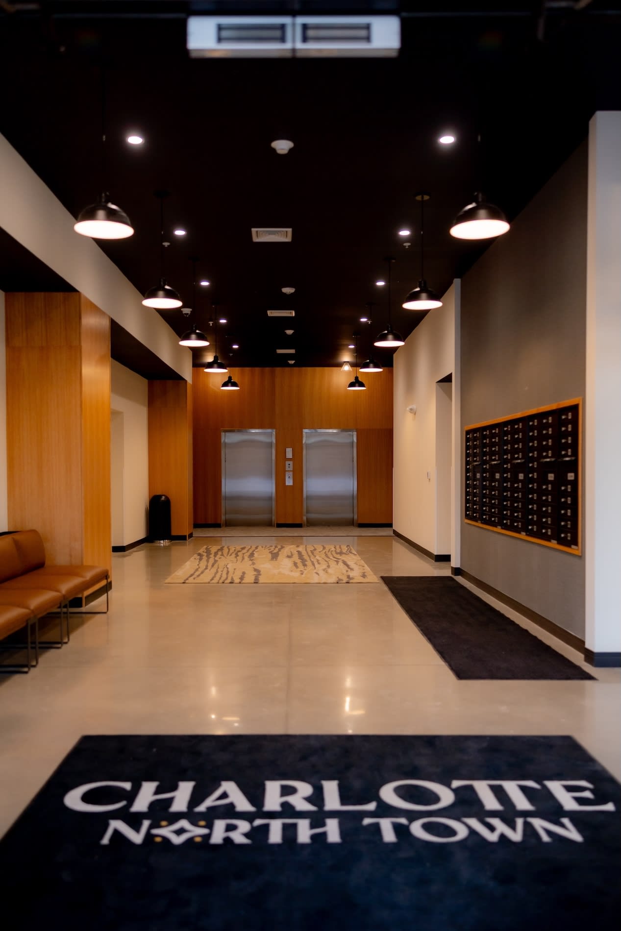 Building lobby with dual elevators, wood-paneled walls, pendant lighting, polished concrete floors, and mailbox wall.