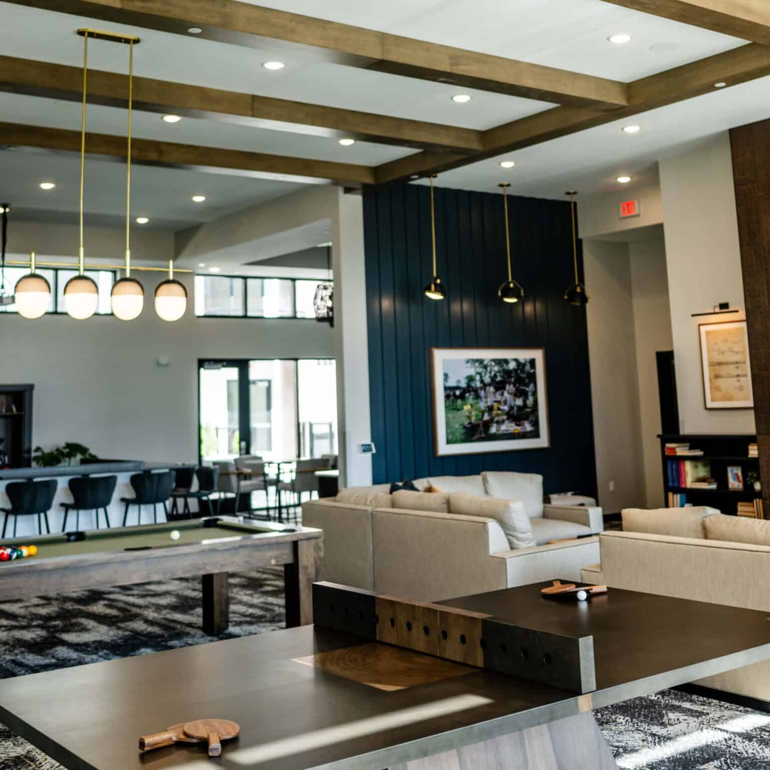 Common room with cream seating, dark blue accent wall, exposed wood ceiling beams, pendant lights, and pool table.
