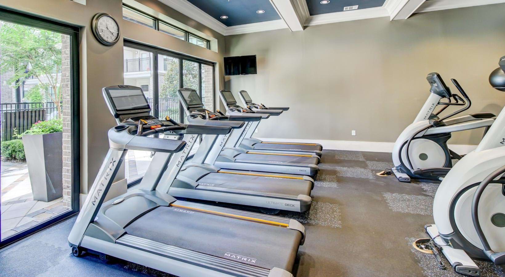 Fitness center with row of treadmills and stationary bikes, large windows overlooking outdoor patio, recessed lighting, and wall-mounted TV.