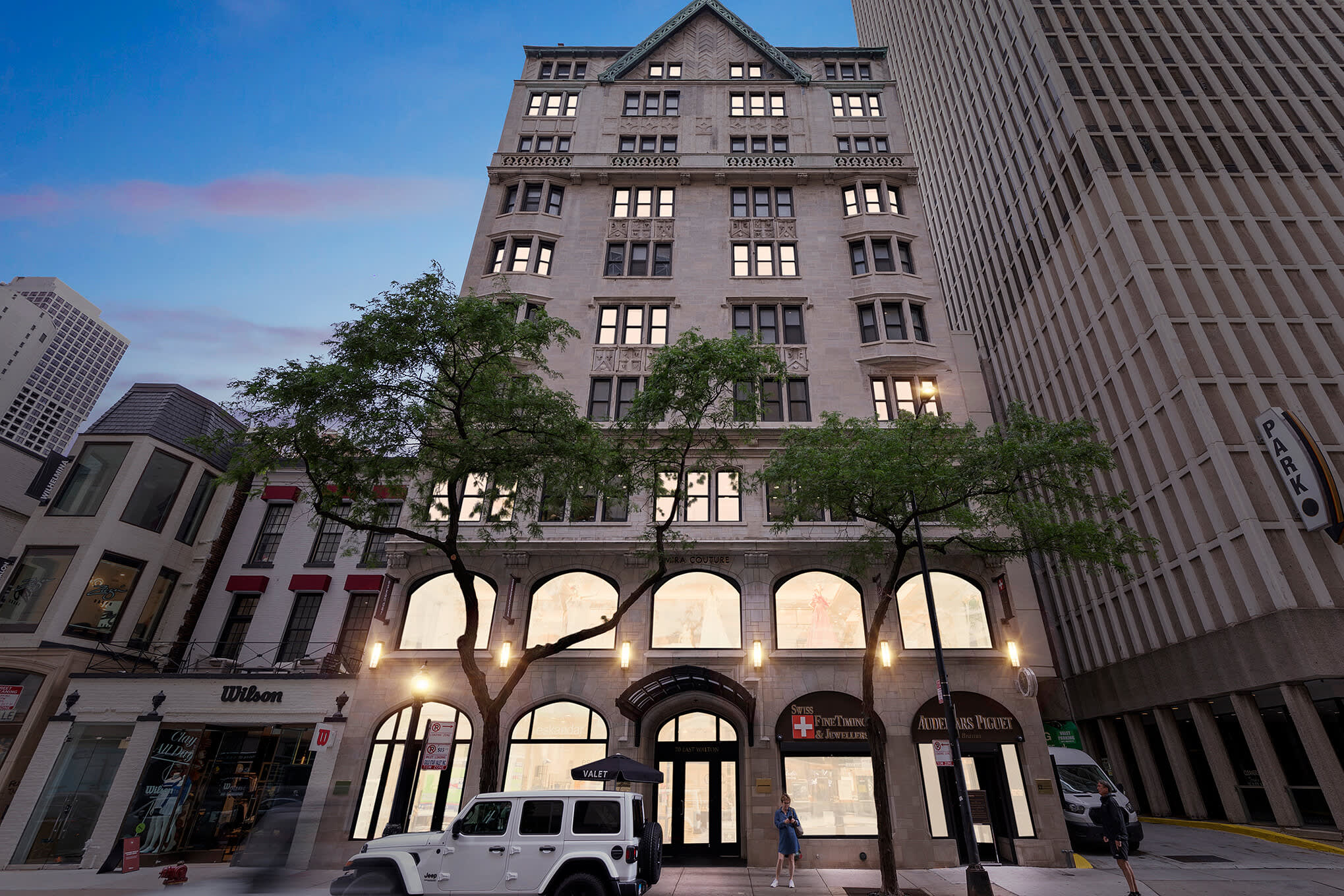 Historic gray stone building facade with arched ground-floor windows, upper-floor residential windows, and mature trees in front.