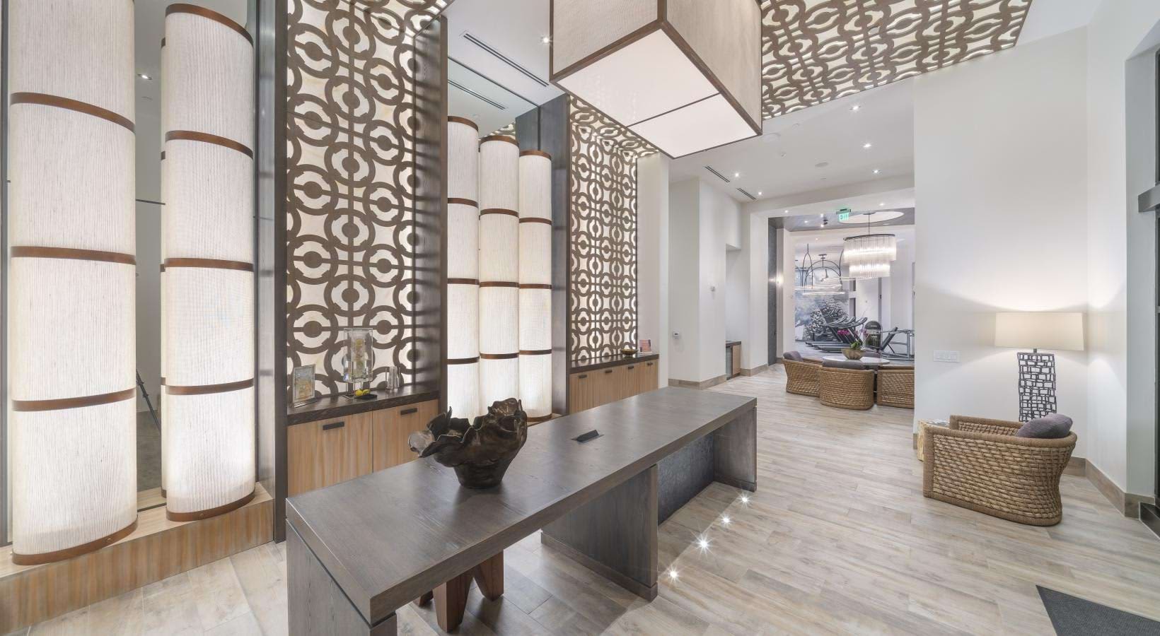 Contemporary lobby with dark wood geometric room dividers, cylindrical pendant lights, concrete desk, and woven seating areas on light wood flooring.