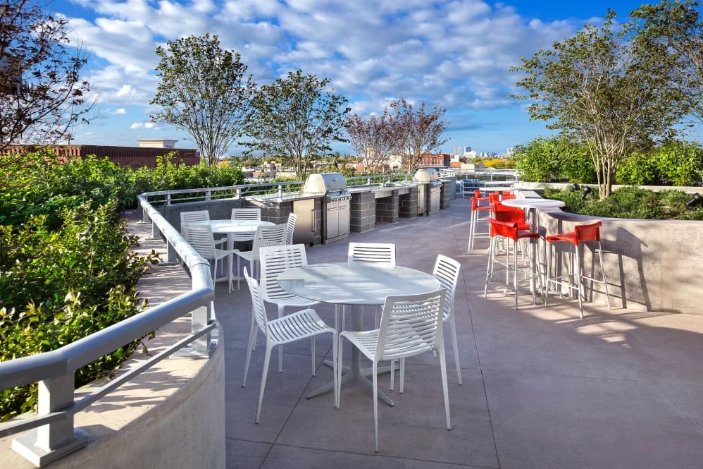 Rooftop terrace with white dining tables, red bar seating, built-in grill, green planters, and Chicago skyline view.