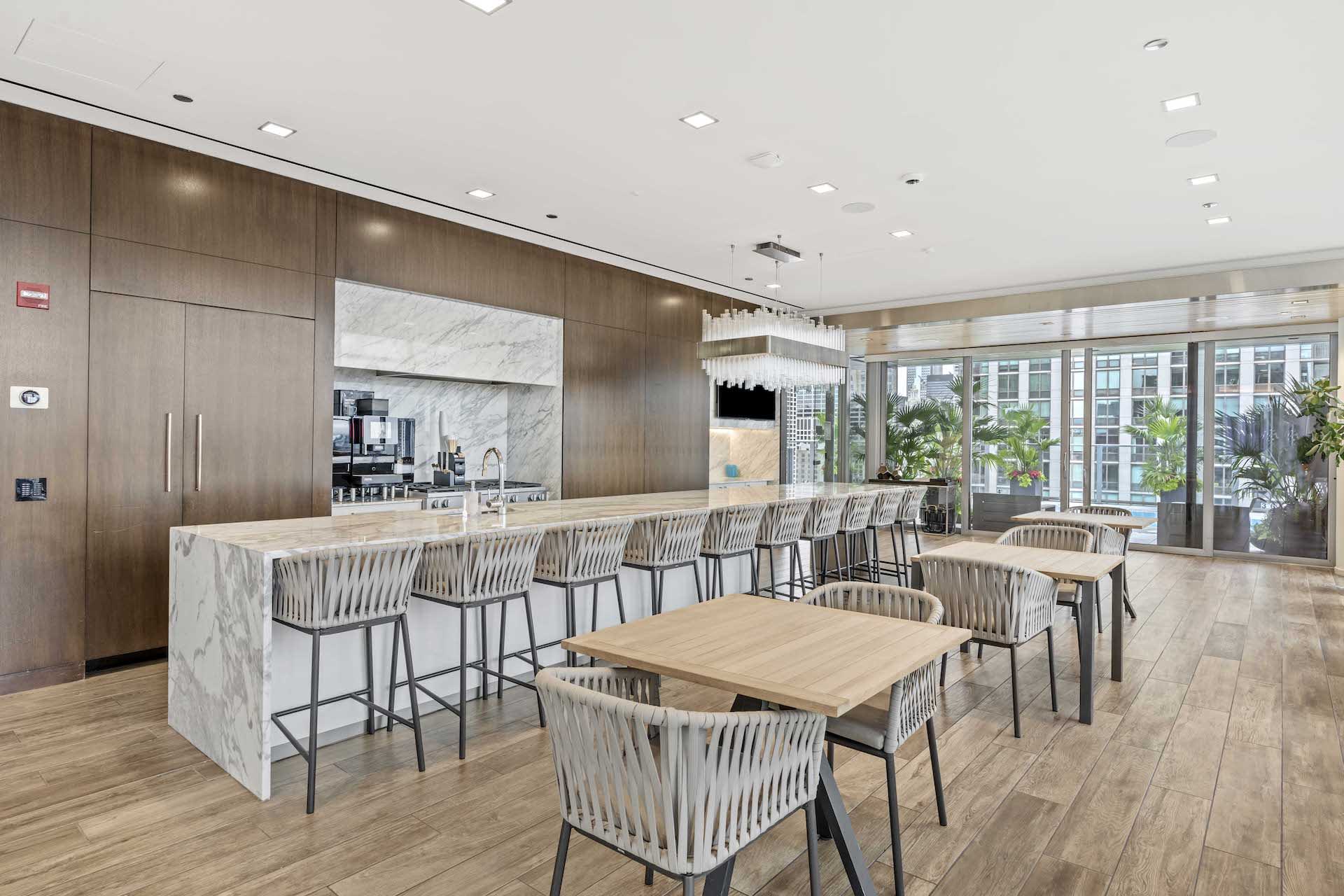 Open-concept kitchen and dining area with marble-topped island, wood cabinetry, woven chairs, and large windows overlooking the city.