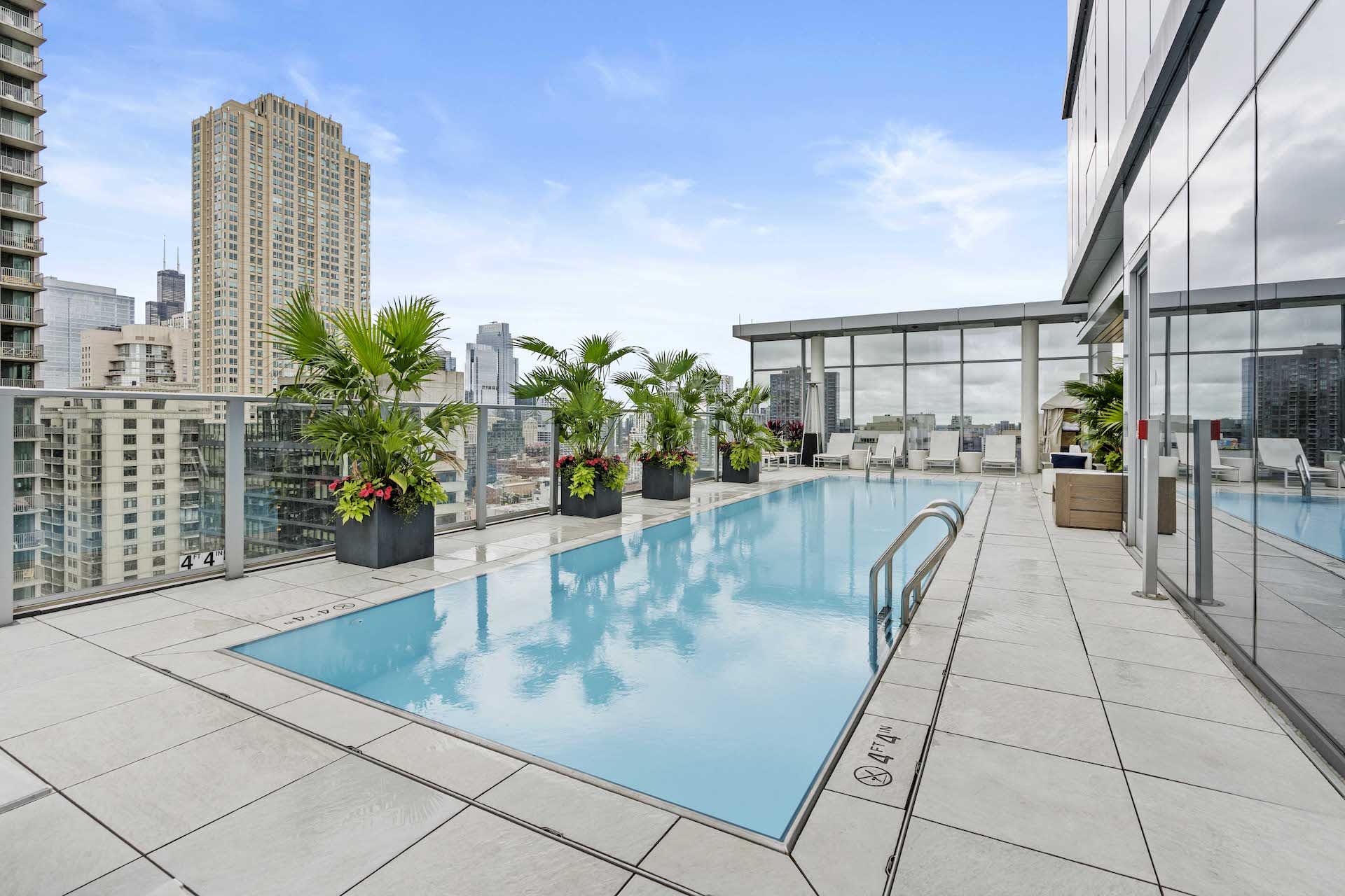 Rooftop pool with clear blue water, potted palm plants, lounge seating, and Chicago skyline views from high-rise building.