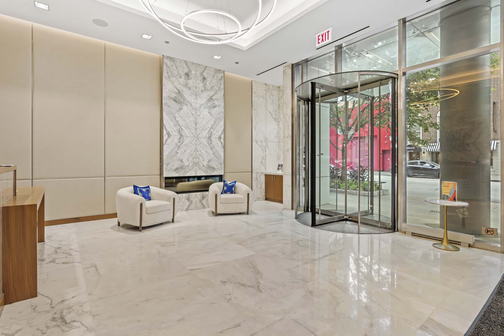 Modern building lobby with white marble floors, cream armchairs with blue pillows, marble accent wall, and glass entrance doors.