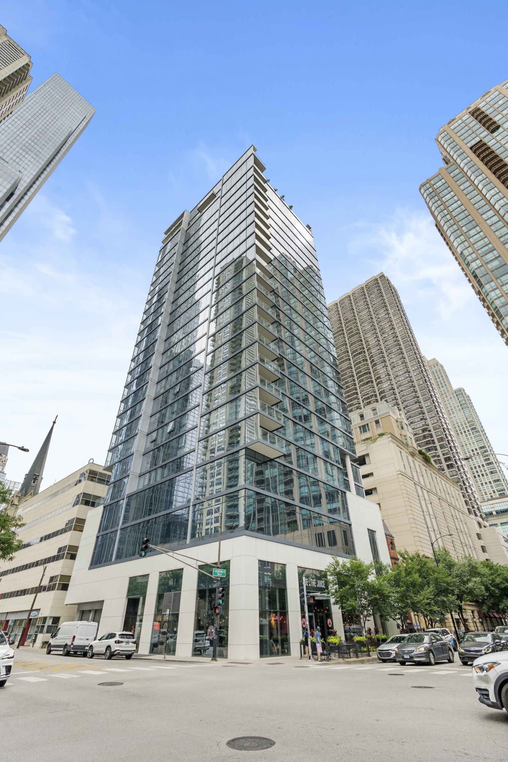 Modern glass and steel residential tower with white base and retail storefronts, surrounded by Chicago skyscrapers and street-level activity.
