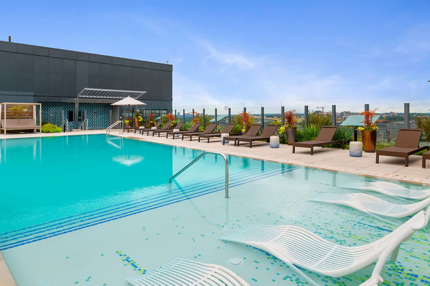 Rooftop pool and spa area with turquoise water, lounge chairs, potted plants, and city skyline views.