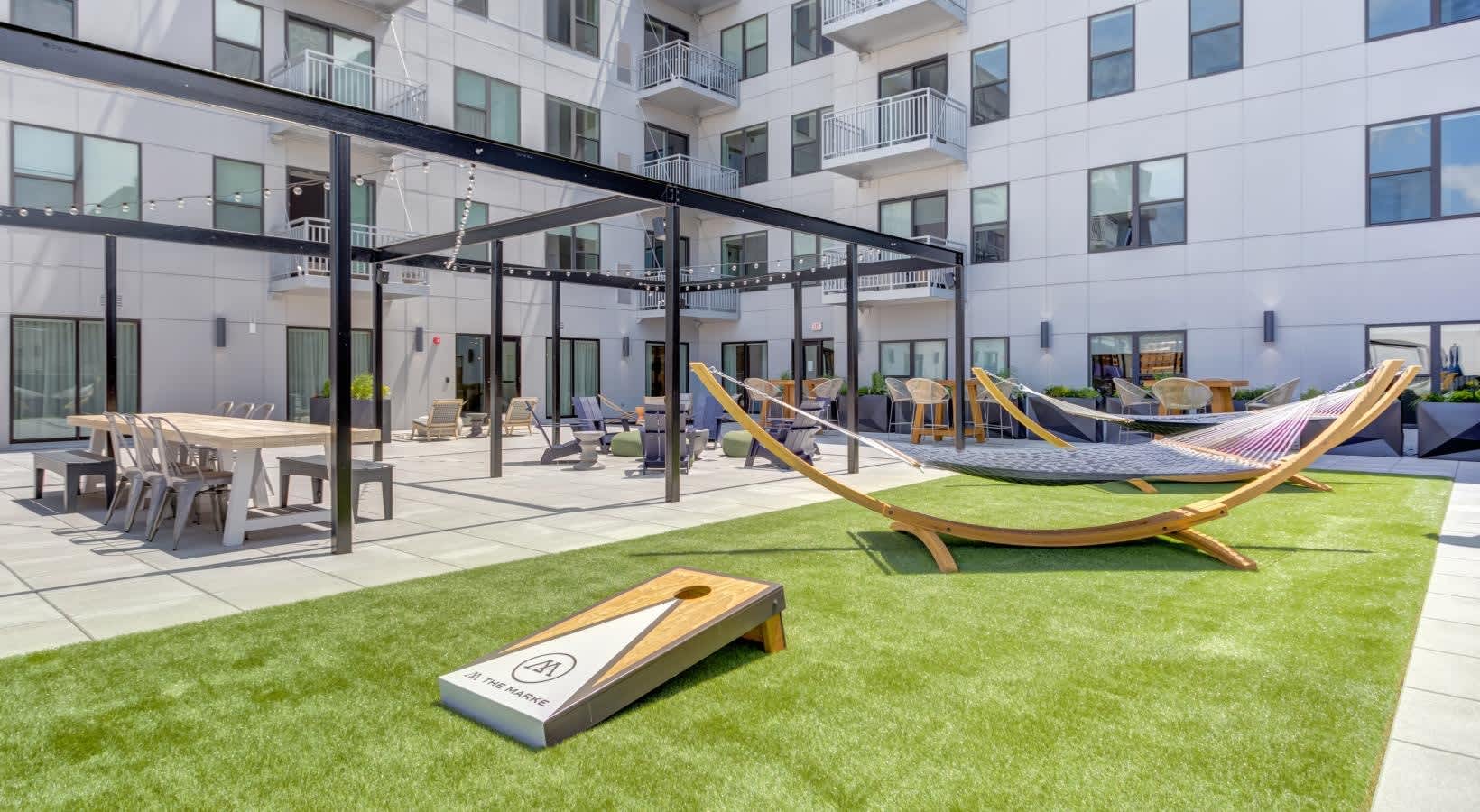 Outdoor courtyard with green lawn, cornhole game, wooden hammocks on stands, black pergola, dining tables, and multi-story white apartment building.