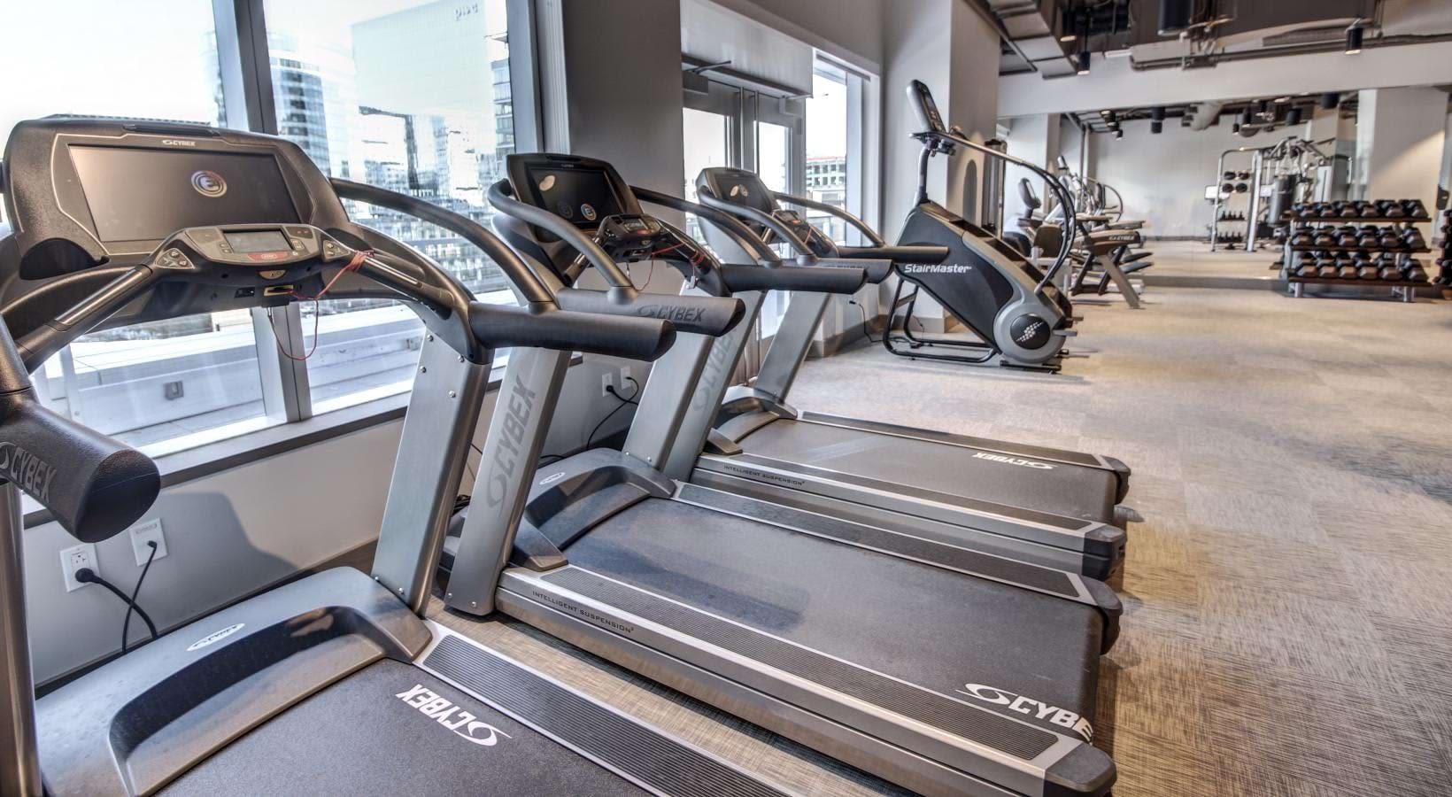 Modern gym with row of treadmills facing windows overlooking Boston skyline, dumbbells and exercise bikes visible in background.