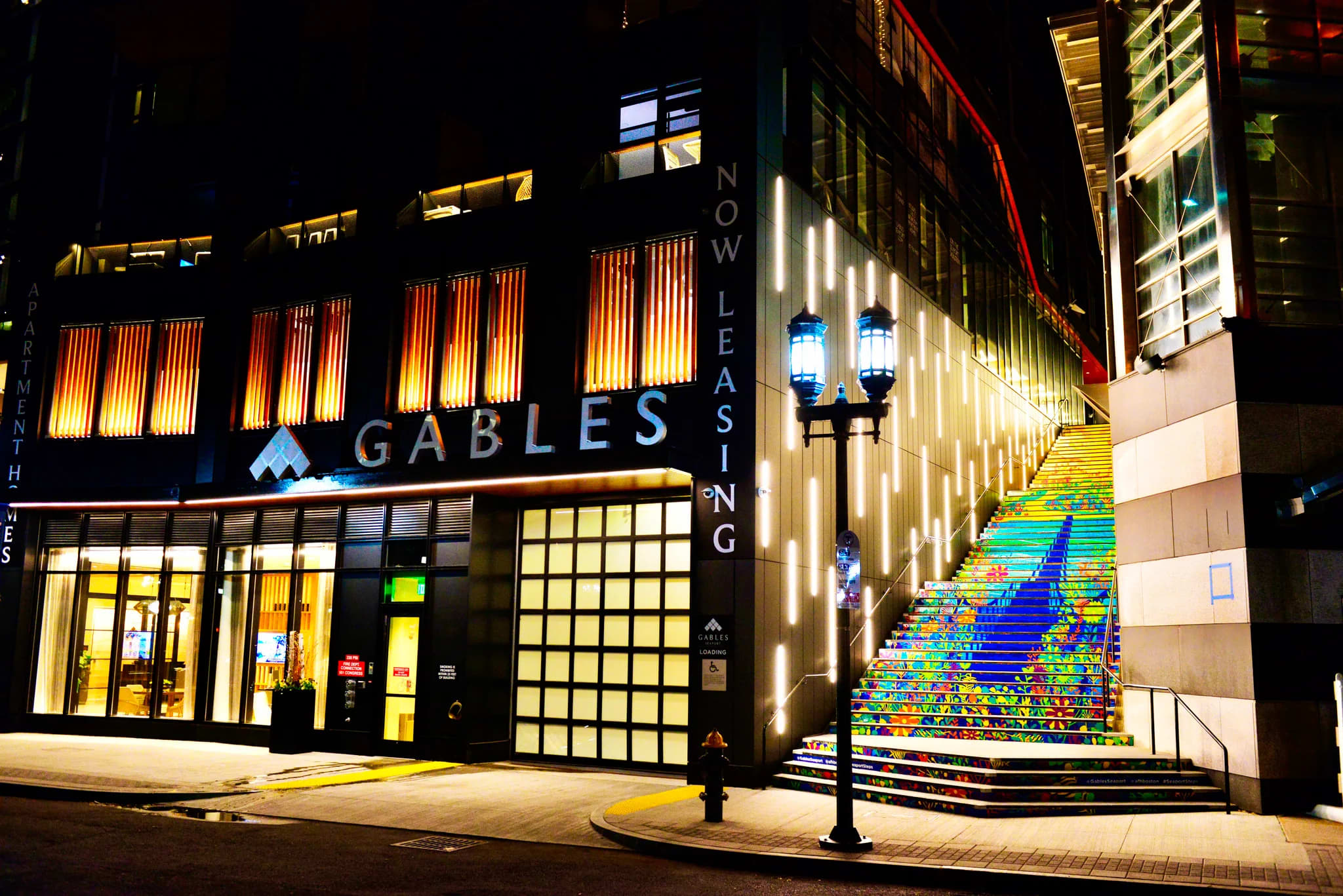 Nighttime exterior of Gables Seaport building with illuminated storefront, adjacent to rainbow-painted public stairs and street lamps.