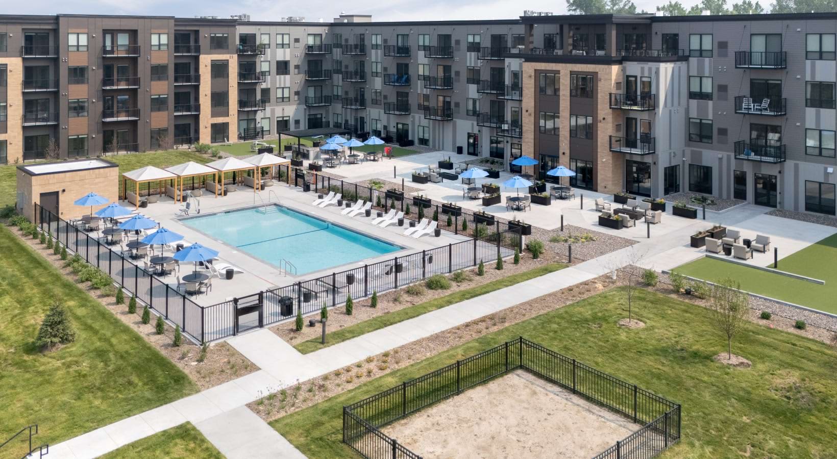 Aerial view of rectangular swimming pool with blue umbrellas and lounge chairs, surrounded by multi-story apartment buildings and manicured grounds.