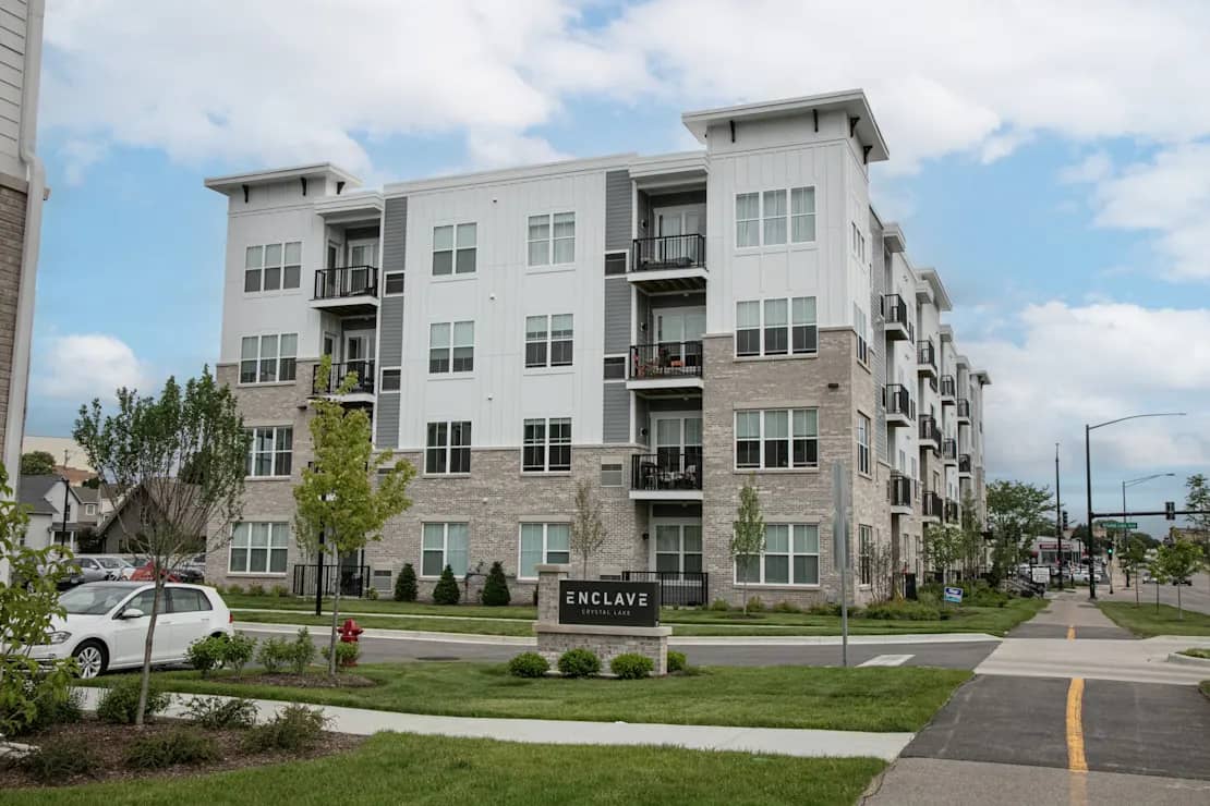 Four-story apartment building with white and brick facade, multiple balconies, and green landscaping in front.