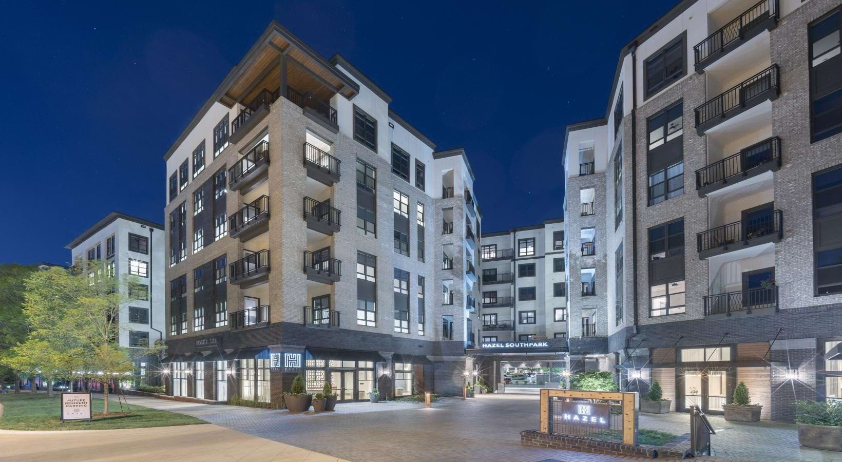 Evening exterior view of contemporary apartment buildings with illuminated windows, brick and stone facades, ground-level retail spaces, and landscaped courtyard.