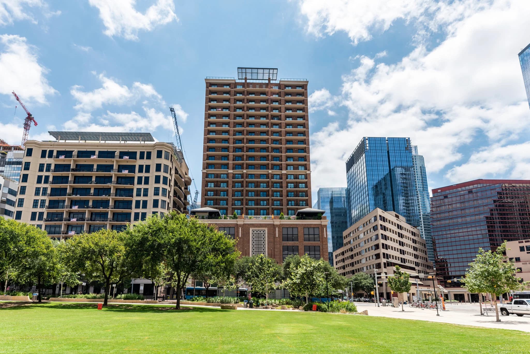 Downtown Austin skyline with mid-rise residential and office buildings surrounding a green plaza with mature trees.