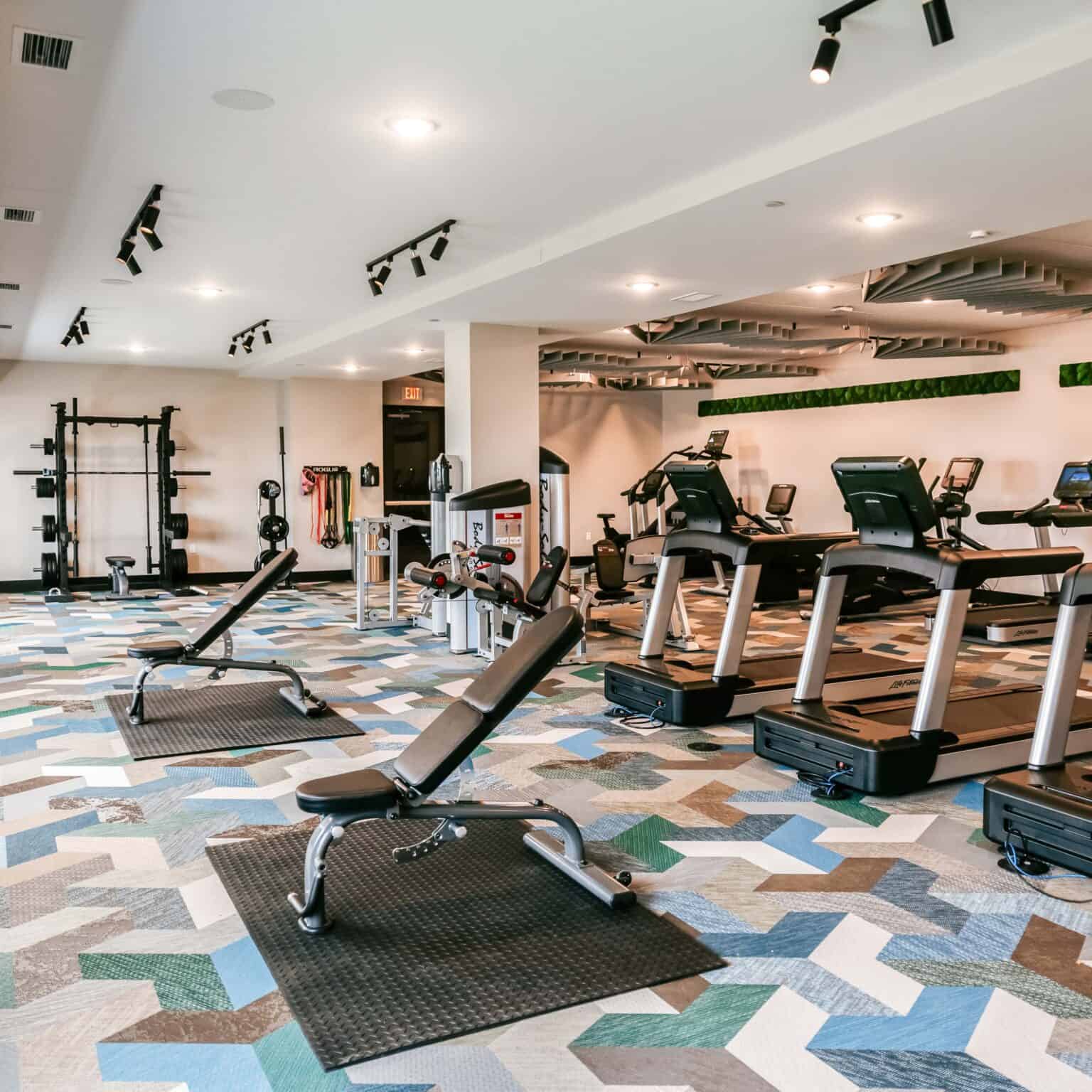 Fitness center with treadmills, exercise bikes, weight benches, and a squat rack on geometric patterned flooring under bright ceiling lights.