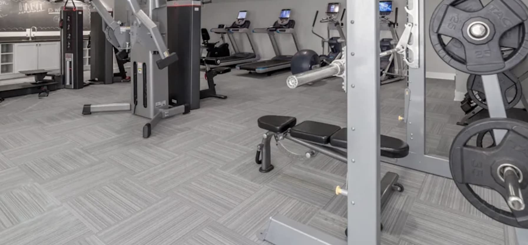 Modern gym with treadmills, exercise bike, weight plates, and strength training machines on gray tile flooring.