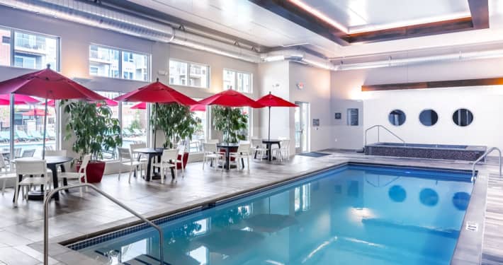 Indoor pool with blue water, red umbrellas, dining tables, and floor-to-ceiling windows in a bright, modern facility.
