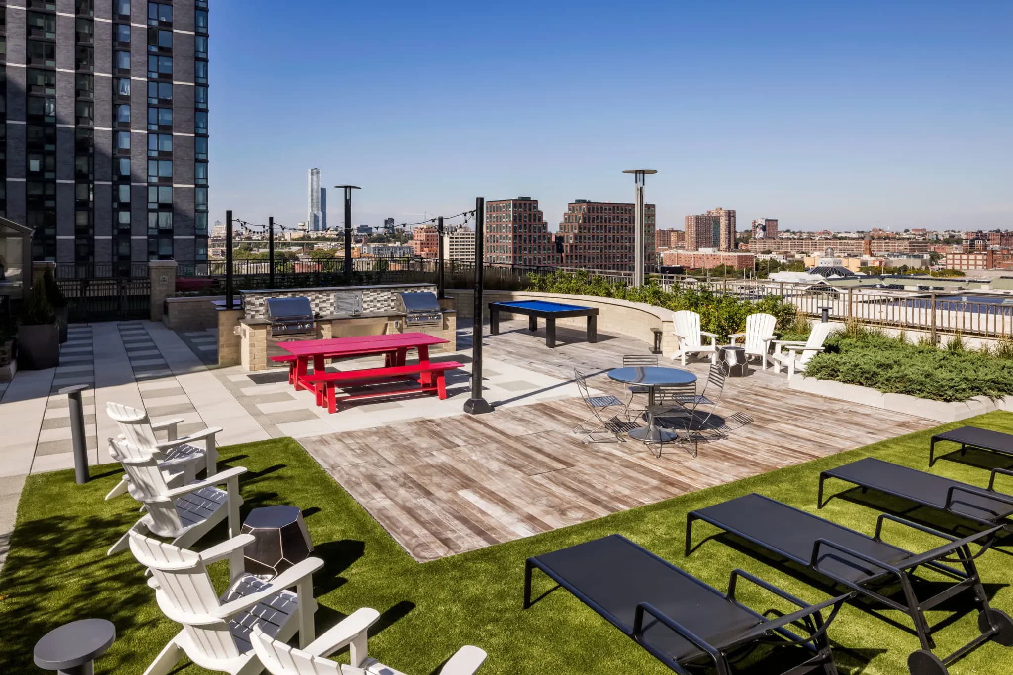 Rooftop terrace with white Adirondack chairs, black loungers, red picnic table, cornhole, and ping pong table overlooking Jersey City skyline.