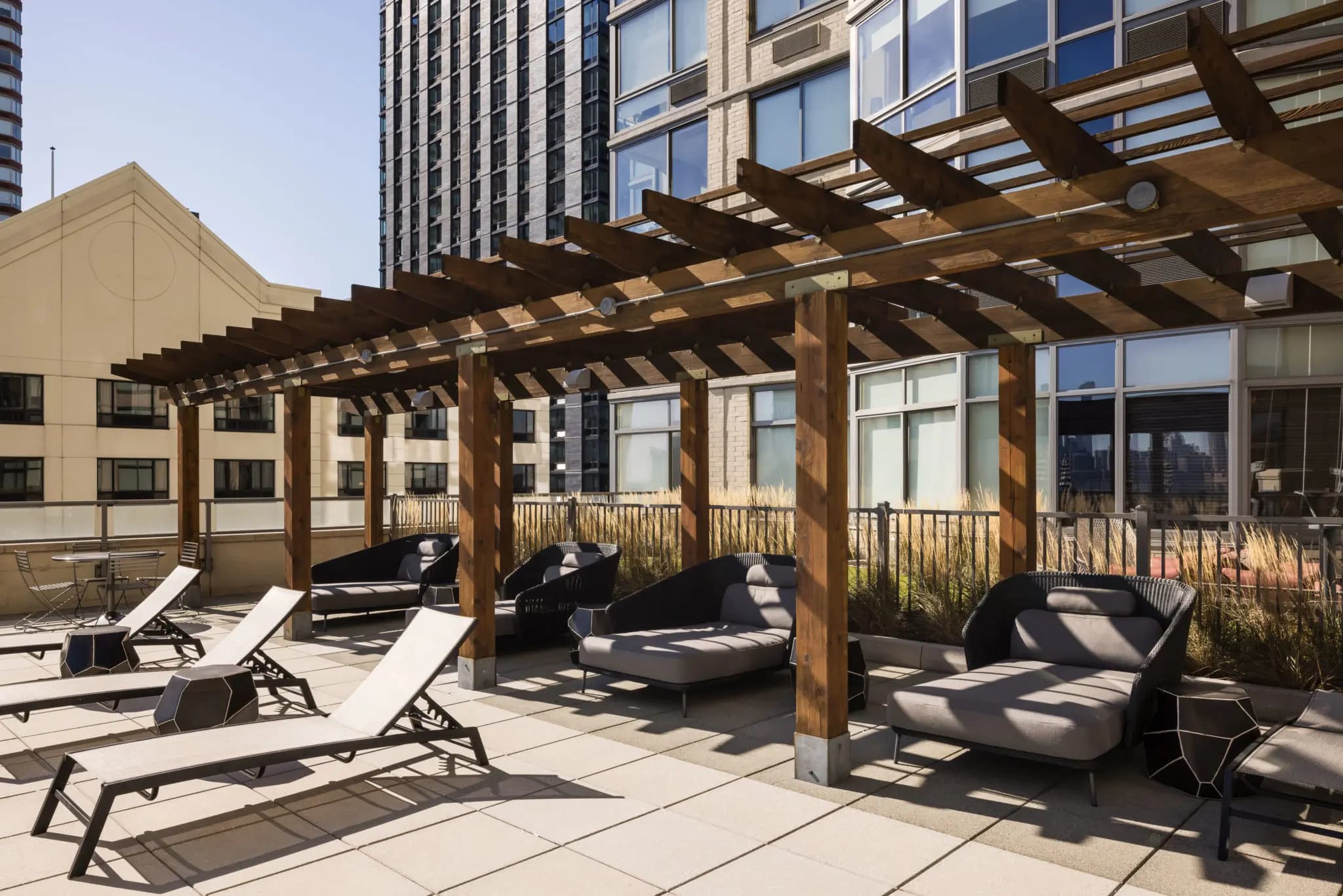 Rooftop terrace with wooden pergola structure, black lounge chairs, and surrounding high-rise buildings in Jersey City.