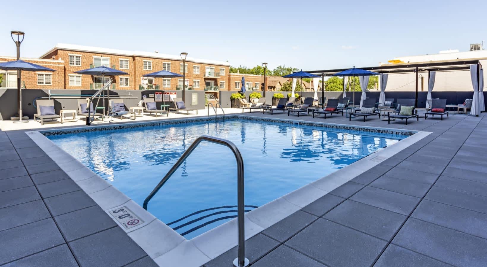 Outdoor swimming pool with black handrail, surrounded by lounge chairs, blue umbrellas, and brick apartment building in background.