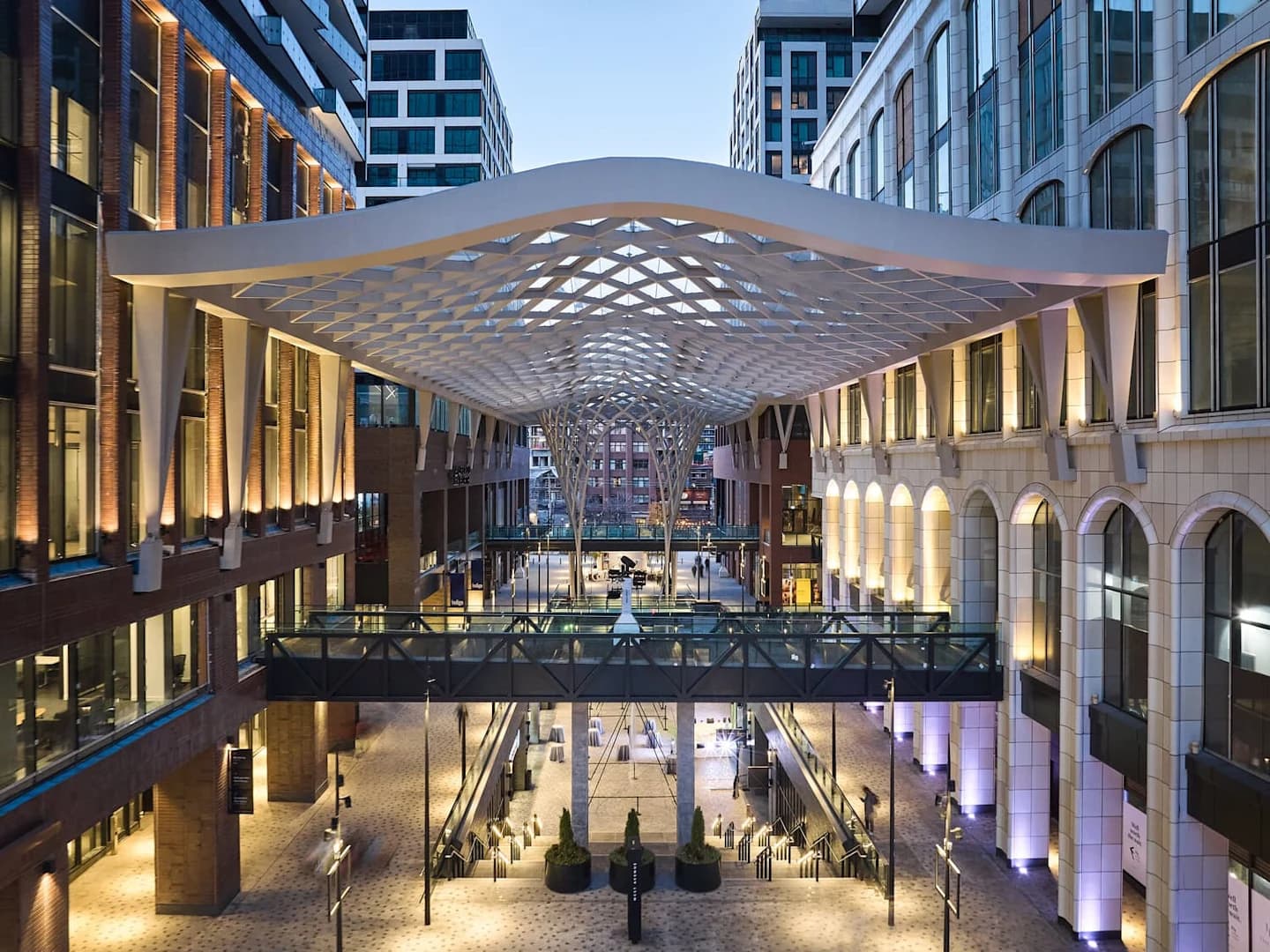 Modern covered courtyard with curved white lattice canopy, illuminated brick buildings on both sides, pedestrian bridge, and stairs leading to plaza below.