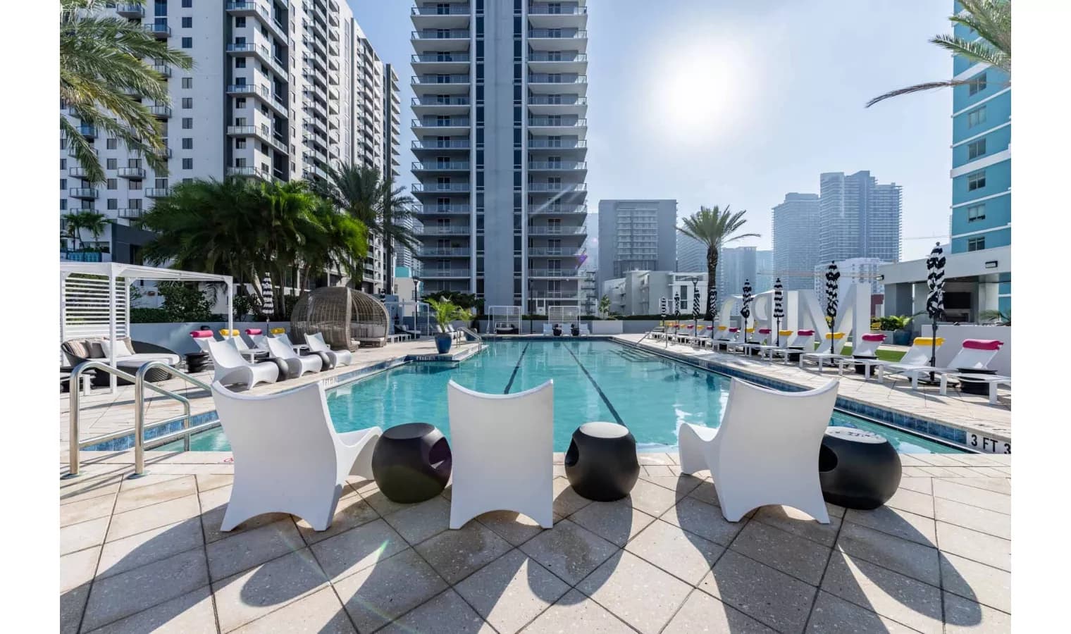 Resort pool with lap lanes, white lounge chairs, black accent tables, and downtown Miami skyline beyond residential towers.