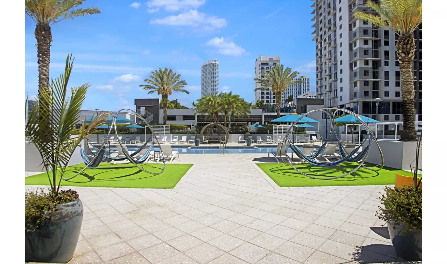 Pool deck with sculptural metal art installations, palm trees, lounge chairs, and high-rise buildings in background.