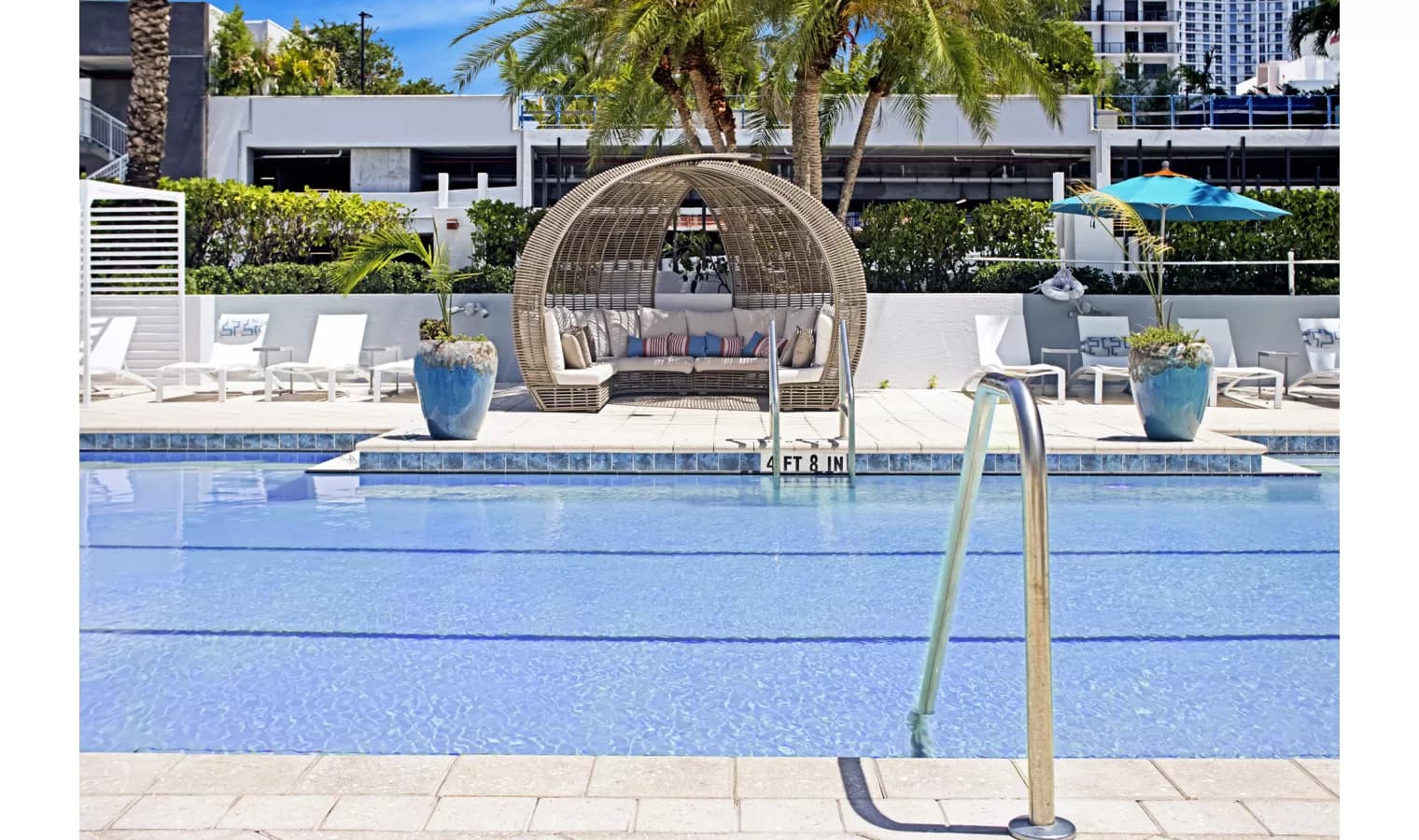 Resort pool with clear blue water, woven egg-shaped lounge pod, palm trees, and white deck chairs under umbrellas.