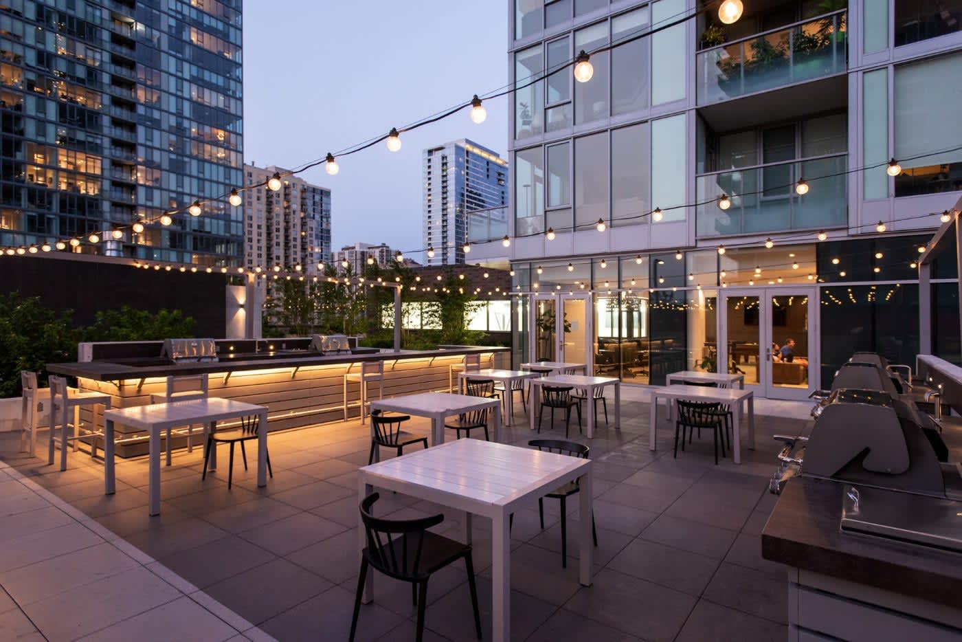 Rooftop terrace at dusk with dining tables, bar counter, string lights, and downtown Chicago skyline in background.
