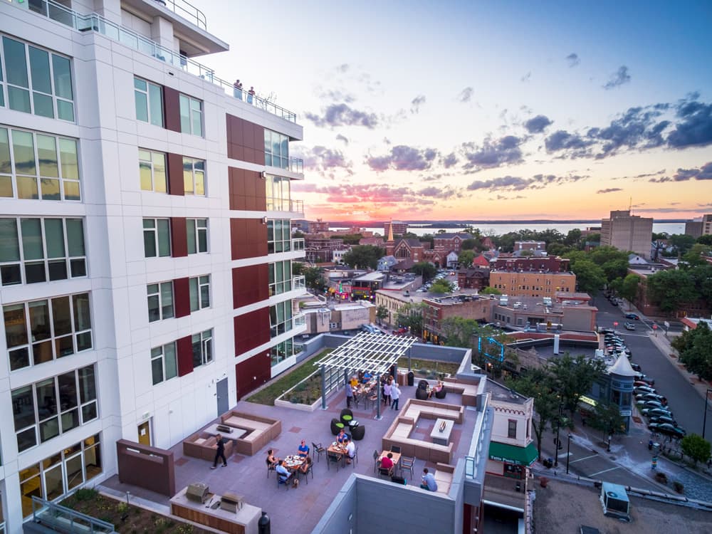 Modern residential building with white and burgundy facade overlooking a courtyard with seating areas, trees, and a sunset view of Madison's downtown and waterfront.